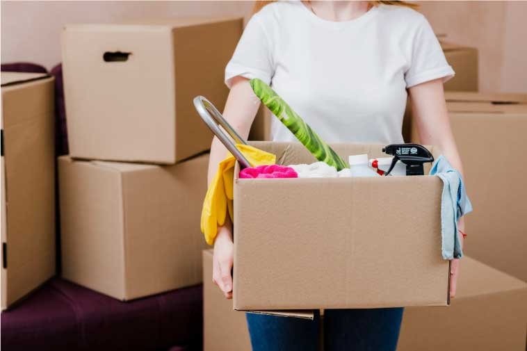 Woman carrying a box with kitchen utensils and moving boxes behind her.